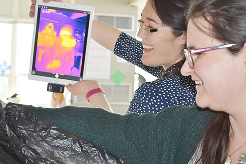 Virginia Lorenz and Pamela Pena Martin do an activity using infrared light. Lorenz is holding an i-Pad loaded with software that uses infrared light to detect the heat signature of a Franklin student who is hidden behind a black garbage bag, holding up several fingers, which Lorenz and Pena Martin are to guess using the infrared image.