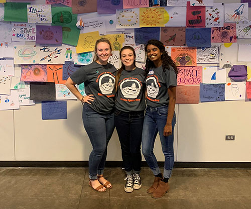  Elena Kamis, Angelina Boynton, and Shivani Ganesh, members of ISS's Educational Outreach Board
 at the fall 2019 Illinois Space Day hosted by ISS. In the background is some of the space artwork completed by young visitors to Illinois Space Day.